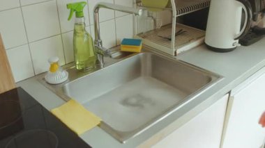 Bir kadın lavaboya sabunlu suyla yaklaşır ve su tesisatı sorununu çözmeye hazırlanır. Temizlik malzemeleri hazır. Işıl ışıl, temiz mutfak..