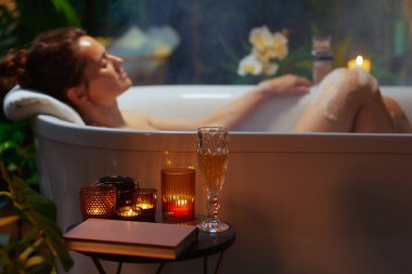 Gözleri kapalı bir kadın dumanı tüten beyaz bir küvette huzur bulur. Kafası havlunun üzerinde dinlenir. Çevredeki mum ışığı, bir kitap ve bir içecek huzur dolu bir gece için lüks bir atmosfer yaratır..