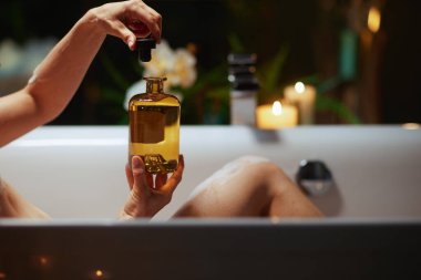 Küvette yakın plan bir el, özenle bir kehribar cam şişeyi açıyor, muhtemelen gerekli yağı içeriyor. Sıcak mum ışığı ve bulanık bitkiler barışçıl, lüks bir self-bakım atmosferi yaratır..