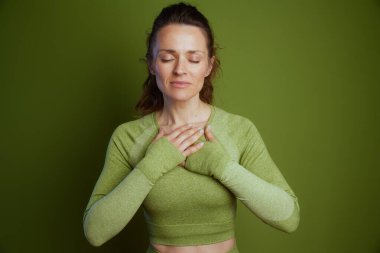 Saçları geriye çekilmiş ve elleri göğsünde açık yeşil atletik bluzlu bir kadın yeşil bir zemine karşı huzurlu bir rahatlama buluyor..