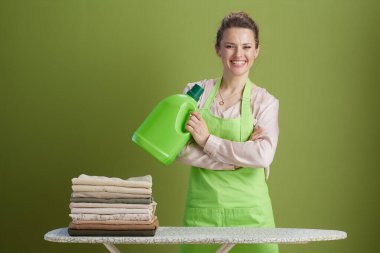 Pembe bluzlu ve yeşil önlüklü gülümseyen bir kadın, düzgün katlanmış nötr renkli elbiseleri olan bir ütü masasının yanında deterjan tutuyor..