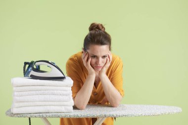 Yorgun bir kadın, evde duygusal yorgunluk gösteren çamaşır ve ütünün yanında elleri üzerinde dinleniyor..