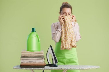 Yüzüne yeni yıkanmış giysiler tutarak gülümseyen bir kadın. Pembe gömlek, yeşil önlük, deterjanlı ütü masası ve katlanmış giysiler açık yeşil arka plana karşı..