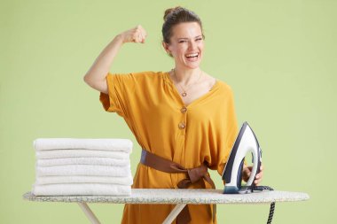 Hardal elbiseli heyecanlı kadın demir ve havluların yanında poz veriyor, ev hayatına olan güvenini ve gücünü ifade ediyor..