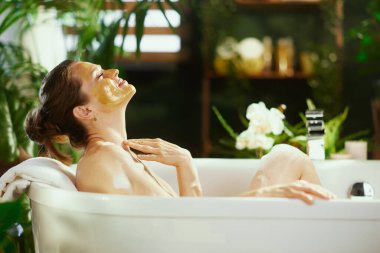 Sarı maskeli bir kadının sakin bir yan profili köpük banyosunda gözleri kapalı rahatlar. Yemyeşil bitkiler ve hafif ışıklandırma, özgüven ve refah için huzurlu bir spa ortamı yaratır..