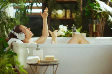 Yetişkin bir kadın kendini neşeyle köpük banyosunda, gözleri kapalı, ağzı açık, kolları havada, kulaklık takarak ifade eder. Lüks, bitkilerle dolu banyo rahatlamak için dingin bir vaha yaratıyor..