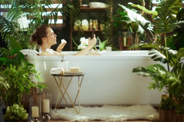 Bir kadın, yemyeşil bitkilerin arasında bir baloncuk ve akıllı telefon tutarak modern köpük banyosundan zevk alır. Sükunet, spa benzeri banyo, yumuşak doğal ışıkla yıkanmış, özbakım ve rahatlamayı somutlaştırıyor..