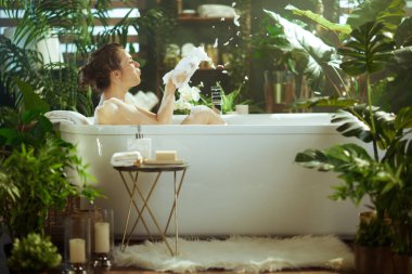 Profildeki bir kadın, beyaz bir köpük banyosunda gevşetiyor, ellerinden köpük saçıyor, yemyeşil bitkiler ve lüks bir spa merkezinde doğal bir ışıkla çevrili..