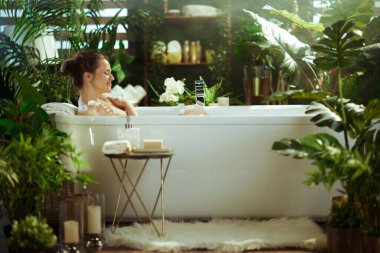 Saçları topuz şeklinde olan bir kadın, huzurlu bir köpük banyosundan, gözleri kapalı, ılık güneş ışığıyla yıkanmış, yemyeşil bitkiler ve sakin bir banyoda spa ürünleriyle çevrili bir şekilde keyif alır..
