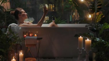 Serbest bir küvette rahatlayan bir kadın akıllı telefonuyla selfie çekiyor. Banyo yeşillik, ışıklar ve mumlarla süslenmiş. Lüks bir atmosfer yaratıyor..