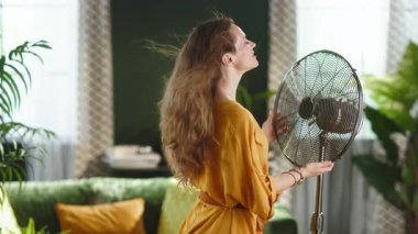 Sarı elbiseli, uzun kahverengi saçlı bir kadın, güneşli bir oturma odasında, eski bir elektrikli vantilatörün serinletici esintisinden zevk alıyor..