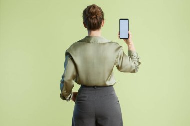 Bir kadın boş bir akıllı telefonu olan seyirciye arkası dönük duruyor. Yeşil bluz ve gri pantolon profesyonel bir bağlam olduğunu gösteriyor..