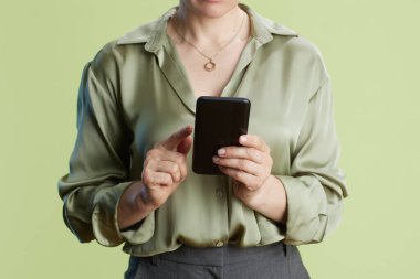 Bir kadın akıllı telefonunu dikkatlice kullanır, parmakları yavaşça ekrana dokunur. Açık yeşil bir bluz ve gri bir etek giymiş açık yeşil bir arka fona karşı.