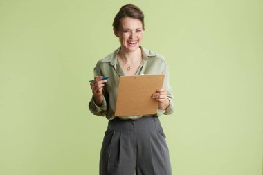 Gülümseyen bir kadın elinde bir not panosu tutuyor, pozitif enerji yayıyor. Yeşil bluz ve gri pantolonla, iş için özgüven ve hazırlık saçar..
