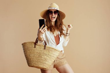 Plaj tatili Gülümseyen 40 yaşındaki beyaz bluz ve şortla bej arka planda hasır çanta, hasır şapka ve cep telefonuyla selfie çeken güneş gözlüklü modern ev kadını..