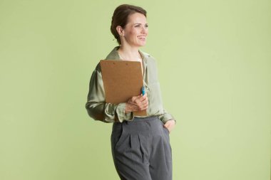 Orta yaşlı bir kadın, bir eli cebinde, elinde bir panoyla gülümsüyor. Açık yeşil bir bluz ve açık yeşil bir arka plana karşı gri pantolon giyer..