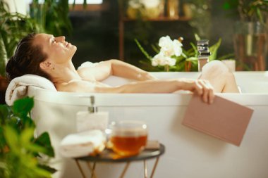 Gülümseyen genç bir kadın, gözleri kapalı, sakin bir köpük banyosunun keyfini çıkarıyor, elinde bir kitapla havluya yaslanıyor. Buhar ve yemyeşil bitkiler beyaz orkidelerle mükemmel bir spa ortamı yaratır.