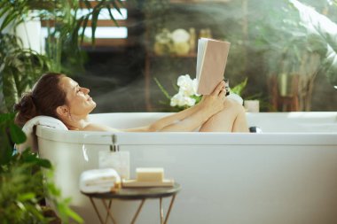 Gözleri kapalı sakin bir kadın beyaz bir küvette kitap okurken hafifçe gülümsüyor. Yumuşak güneş ışığı, buharlı sis ve yemyeşil bitkilerle yıkanmış, rahatlama ve kendine bakımı somutlaştırıyor..