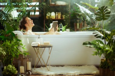 Bir kadın lüks bir köpük banyosunda neşeyle gülüyor, tablet tutarken yukarı bakıyor. Bereketli bitkiler ve puslu atmosferle çevrili bu parlak, spa benzeri banyo, saf özbakım ve neşe sunar..