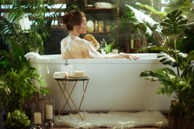 Arkadan, bir kadın sırtını yemyeşil bitkiler ve yumuşak buharla çevrili köpüklü bir banyoda ovalıyor. Bu sakin sahne, modern bir kaplıcada kişisel bakım, rahatlama ve sağIığı yakalar..