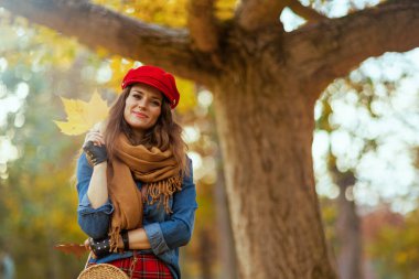 Merhaba sonbahar. Şık kadın portresi kot gömlek ve sonbahar yaprağı, atkı ve eldivenli kırmızı şapka şehir parkındaki ağacın yanında..