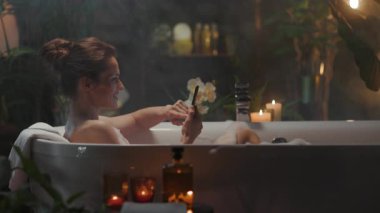 Bun saç stiline sahip rahat bir kadın, akıllı telefonuna bakarken köpük banyosu yaparak modern bir özbakım anından zevk alır. Sakin, spa benzeri banyo, bitkiler ve mumlarla süslenmiş, dijital eğlenceyi sükunetle harmanlıyor..