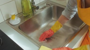 Turuncu önlüklü bir insan paslanmaz çelik bir mutfak lavabosunu sabunlu suyla temizler. Sürekli tıkanma ve su tesisatına ihtiyaç olduğunu ima eder..