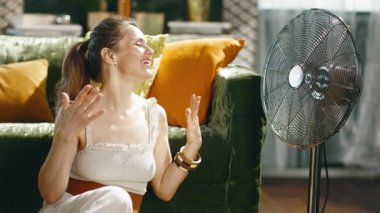 At kuyruklu bir kadın, gözleri kapalı ve gülümsüyor, oturma odasında yerde dinleniyor, sıcak, güneşli bir günde ayakta duran vantilatörün serinletici esintisinin tadını çıkarıyor, rahatlık ve rahatlama iletiyor..