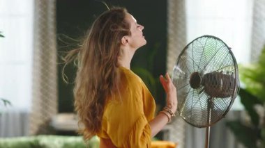 Sarı bir bluz giyen, dalgalı kahverengi saçlı, gülümseyen bir kadın, parlak bir oturma odasında duruyor, ayakta duran bir yelpazenin serinletici esintisinin tadını çıkarıyor, yaz tatili.