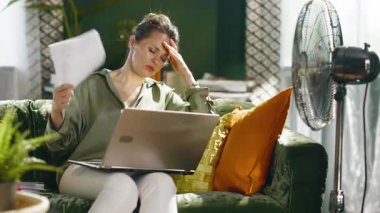 Kadın yeşil kadife bir kanepede dizüstü bilgisayarıyla çalışıyor, yaz sıcağıyla boğuşuyor, kendini kağıtlarla körüklüyor. Sıcak hava dalgasının ev ofis verimliliğini etkilemesinden kaynaklanan rahatsızlık ve hayal kırıklığı.