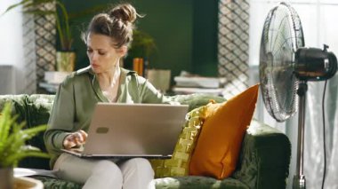 Odaklanmış bir kadın, yeşil kadife bir kanepedeki belgeleri inceleyerek laptopunda etkili bir şekilde çalışıyor. Ayakta duran bir yelpaze, sıcak havalarda rahat ve verimli bir uzaktan çalışma olduğunu belirten hoş bir esinti sağlar..