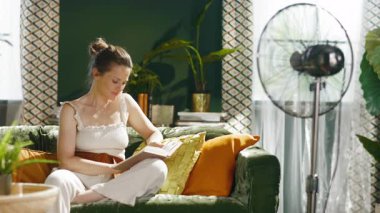 Bir kadın yeşil kadife bir kanepede rahatça rahatlar, mutlu bir gülümsemeyle bakmadan önce kitap okur. Metal zemin pervanesi ve doğal ışık evindeki barışçıl, kendi kendine bakım atmosferini arttırıyor..