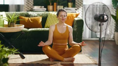 Hardal sarısı spor kıyafetli sakin bir kadın meditasyon yapıyor, gözleri kapalı, güneşli bir oturma odasında. Büyük bir metal vantilatörü serinletici bir rahatlık sağlıyor, ev sağlığı rutinini geliştiriyor..