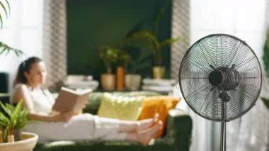 Önünde yelpaze olan genç bir kadın yeşil bir kanepede kendini kitaplara kaptırıyor. Yumuşak doğal ışıkla yıkanan bitki dolu yaşam alanı, rahat bir yaz ortamı yaratır..