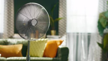 Parlak bir oturma odasının ön planında, renkli yastıklı yeşil bir kanepe, desenli perdeler, ve büyük bir pencere, sakin, rahat bir ev ortamını taşıyan bir gümüş kaide vantilatörü..