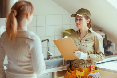 Bronz üniformalı ve beyaz eldivenli bir tesisatçı, parlak bir mutfak ortamında müşterisiyle ev onarımını tartışıyor..