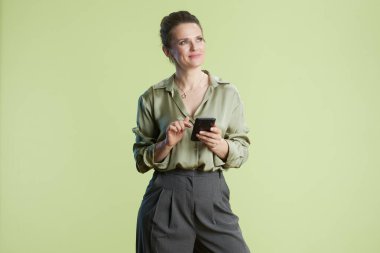 Hafif gülüşlü, yeşil bluz ve gri pantolon giyen akıllı telefonunu kullanan bir kadın. Soluk yeşil arka plan ve yumuşak ışıklandırma modern, düşünceli bir his yaratır..