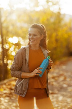 Merhaba sonbahar. Parkta elinde bir şişe suyla spor kıyafetleriyle mutlu, orta yaşlı bir kadın..