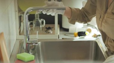 Bronz tulumlu ve beyaz eldivenli tesisatçı, kurulumdan sonra paslanmaz çelik lavabodaki modern krom musluktan akan suyu kontrol ediyor..