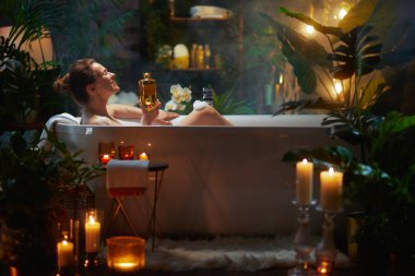 Bir kadın köpük banyosunda, elinde altın bir şişeyle, bitkilerin ve mum ışığının arasında rahatlatıcı ve lüks bir spa deneyimi yaratıyor..