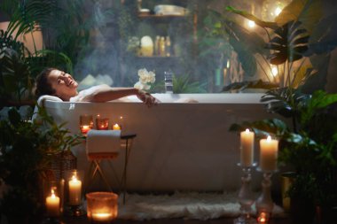 Gülümseyen bir kadın, yemyeşil ve yumuşak mum ışığında köpük banyosunda rahatlar. Huzurlu bir spa ortamı yaratır..