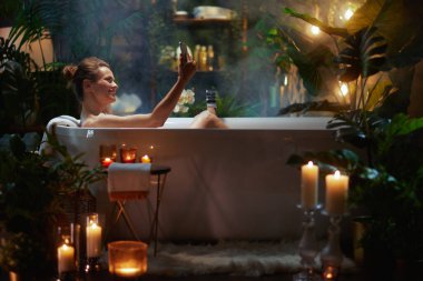 Bir kadın, mum ışığında, bitki dolu bir banyoda cep telefonunu kullanarak köpük banyosunda rahatlıyor. Sahne modern özbakım ve dijital bağlantıyı vurguluyor.