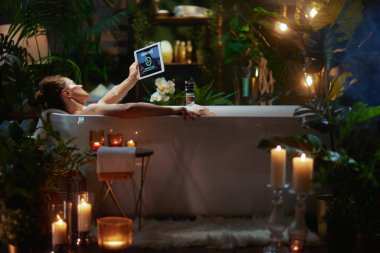 Bir tabletle köpük banyosunda dinlenen bir kadın, 'Favori Film' izliyor. Yeşillik bitkiler ve mumlar, huzurlu, lüks ve özverili bir an yaratır..