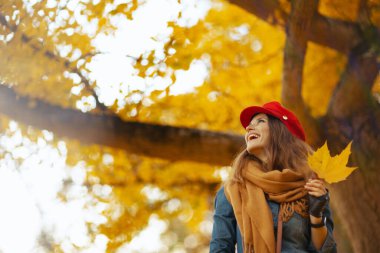 Merhaba sonbahar. Şehir parkında kot gömlekli, kırmızı şapkalı, güz yaprağı, atkısı ve eldivenli mutlu şık bir kadın..
