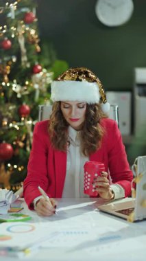 Noel zamanı. Düşünceli, 40 yaşında, Noel Baba şapkalı, kırmızı ceketli, belgeli, kahve ve dizüstü bilgisayarlı, modern yeşil ofiste Noel ağacıyla çalışan bir iş kadını..