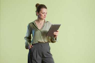 Kendine güvenen bir kadın açık yeşil bir zemine karşı bir tablet tutuyor, modern iş dünyasını, profesyonel hayatı ve teknoloji kullanımının kolaylığını temsil ediyor..