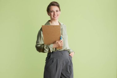 30 'lu yaşlarda gülümseyen bir kadın bir elinde kağıt ve kalem, bir elinde de cebinde. Yeşil bir bluz ve gri pantolon giyiyor. Eşleşen yeşil bir arka planı var..