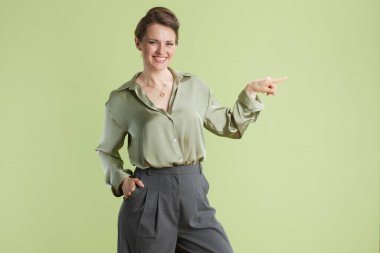 Açık yeşil ipek gömlekli ve gri pantolonlu gülümseyen bir kadın, yeşil zemine karşı güven ve olumlu bir tutum sergileyerek sağı işaret ediyor. Sunumlar için mükemmel.
