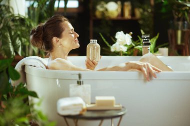 Huzurlu orta yaşlı bir kadın lüks beyaz bir küvette gevşiyor, gözleri huzur içinde kapalı, yemyeşil bitkilerin arasında bir şişe banyo tuzu ve nihai özen için doğal bir ışık tutuyor..