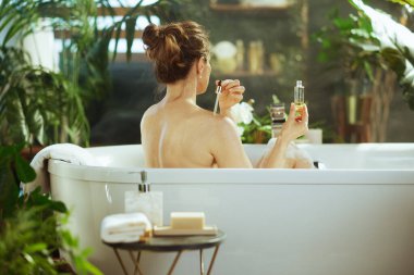 Bir kadın beyaz bir küvette gevşiyor ve omzundan aldığı sıvıyı cilt bakımı için kullanıyor. Bereketli bitkilerle ve doğal ışıkla çevrelenmiş bu dingin spa, rahatlamayı ve refahı teşvik eder..